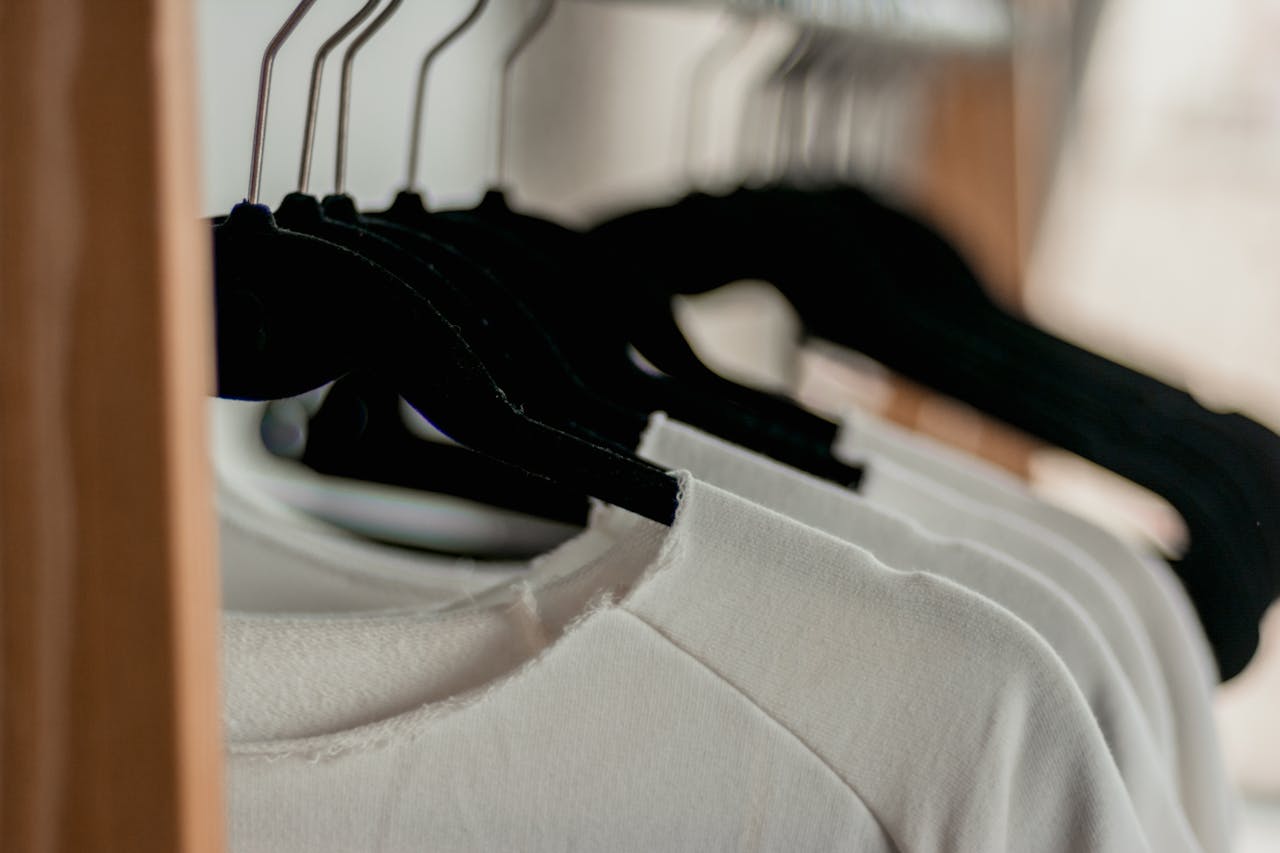 hero-homepage Close-up of white t-shirts hanging neatly on black hangers in a minimalist indoor closet setting.